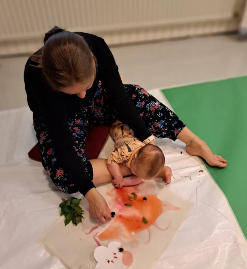 Kuvassa äiti ja vauva. vauva mahaa mahalleen äidin jalan päällä maalausalustalla ja ihmettelee värejä.