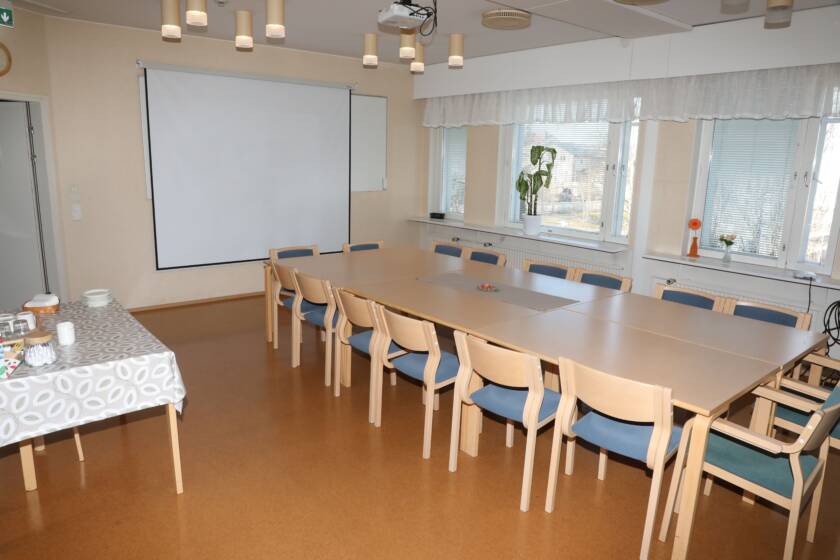 Iso kokouspöytäryhmä, taustalla seinällä valkokangas.