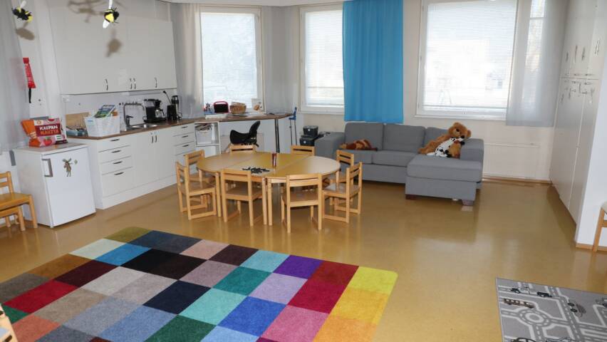 Kerhohuone, jossa on matto, joka on hyvin värikäs, huoneessa on pöytä lapsille sekä harmaa sohva.
