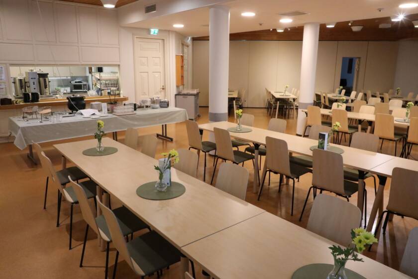 Monta pöytäryhmää tuoleineen seurakuntakeskuksen kahviossa sekä tarjoilupöytä.