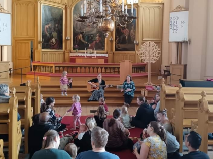 kirkossa edessä kaksi aikuista istumassa kirkon alttarin portailla ja lapsiperheitä istumassa kirkon lattialla ja osallistumassa muskariin.