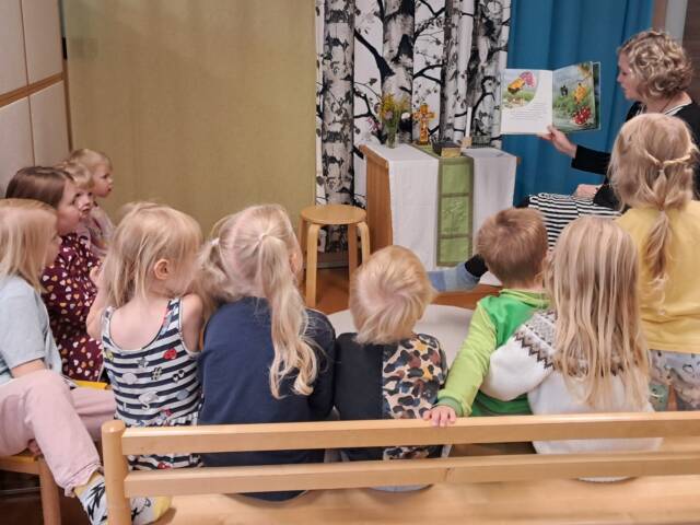 Kuvassa lapsia istumassa penkillä kuuntelemassa kun kerhon ohjaaja lukee kirjaa.