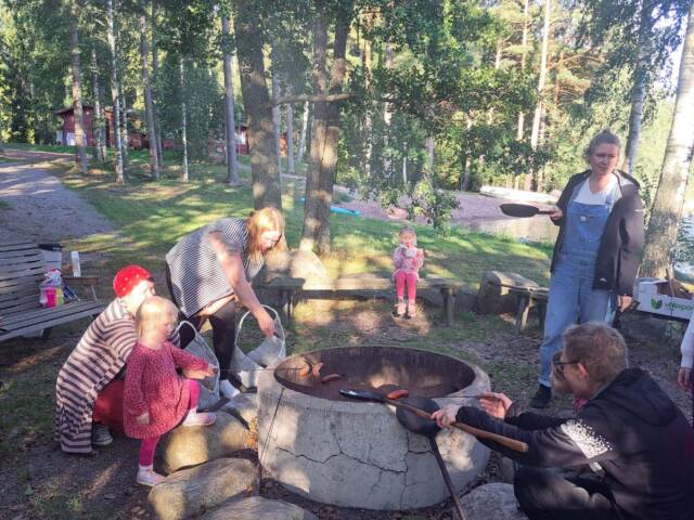 Kuvassa lapsia ja aikuisia grillaamassa makkaraa ja paistamassa lettuja nuotiolla. aurinko paistaa puiden takaa.