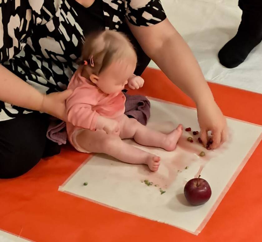 kuvassa istuva vauva ja äiti yhdessä ihmettelemässsä värikylvyssä omenaa ja värinä käytettäviä marjoja.