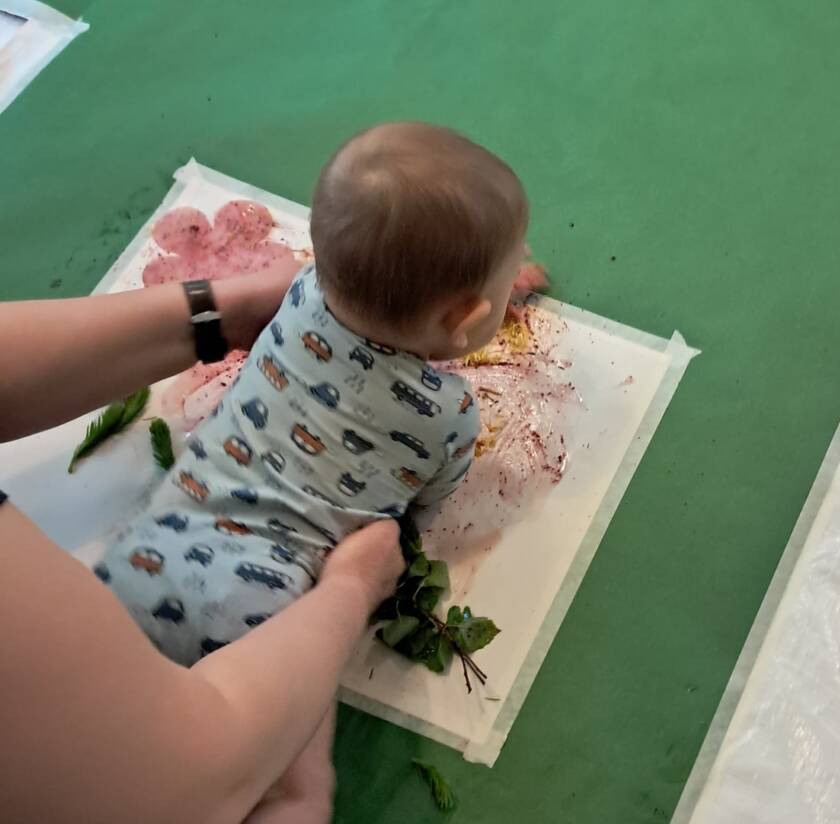 Kuvassa vauva makaa vatsallaan maalausalustan päällä, johon on jo maalattu paljon. kuvassa näkyvät myös kädet, jotka kannattelevat vauvaa kainaloista.
